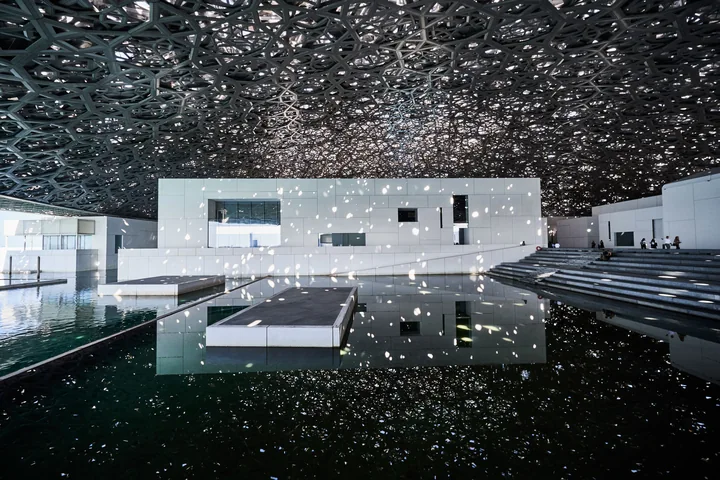Sunlight falling through the patterned dome onto white galleries and water at Louvre Abu Dhabi